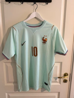 Frankrike vm tröja 2026 - Snygg ljusblå fotbollströja med guldiga detaljer, nummer 10 och Mbappé på ryggen. Tröjan har korta ärmar, broderad fransk tupp och två stjärnor på bröstet. Ränder i blått, vitt och rött vid ärmsluten. Perfekt för dig som älskar fotboll och vill stötta Frankrike.