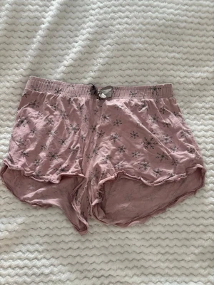 Rosa pyjamasshorts med snöflingor - Söta rosa pyjamasshorts från mywear med grått snöflingemönster och volangkant. Shortsen har resår i midjan och en liten rosett framtill. Perfekta för mysiga hemmakvällar eller sommarens varma nätter.