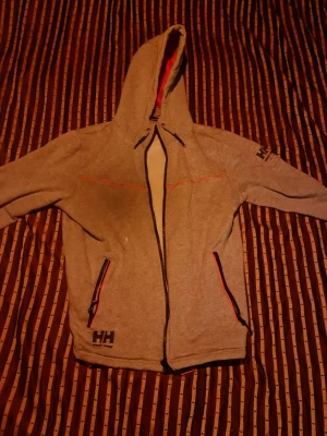 Grå hoodie med dragkedja från Helly Hansen - Snygg grå hoodie från Helly Hansen med dragkedja framtill och två fickor med svarta och röda detaljer. Stor huva och HH-logga på bröstet och ärmen. Perfekt för chill dagar eller när du vill ha en sportig look. Tillverkad i mjukt material.
