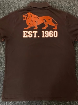 Brun pikétröja från Lonsdale - Den sitter som storlek M. Brun pikétröja från Lonsdale med orange logga på bröstet och stort lejontryck samt texten 'EST. 1960' på ryggen. Klassisk krage och knäppning framtill. Perfekt för en sportig och avslappnad stil.