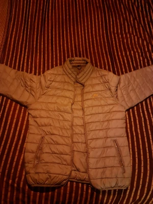 Beige quiltad pufferjacka från Nike - Säljer en beige pufferjacka från Nike med quiltad design och hög krage. Jackan har dragkedja framtill, två fickor med dragkedja och en broderad Nike-logga på bröstet. Perfekt för kyliga dagar och enkel att matcha med olika outfits.