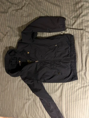 Mörkblå vindjacka med huva - Snygg mörkblå vindjacka från Lyle & Scott med gul logga på bröstet. Jackan har huva, dragkedja framtill och två fickor med tryckknappar. Elastisk nederkant och ärmslut för skön passform. Perfekt för blåsiga dagar.