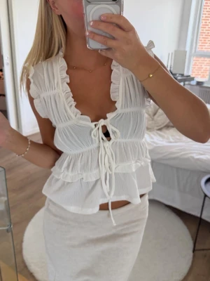 Vit volangblus med snörning - Supersöt vit blus med volangdetaljer och djup v-ringning. Toppen har snörning framtill och är ärmlös, vilket ger en luftig och somrig känsla. Perfekt att styla med kjol eller jeans för en trendig look.