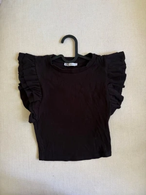 Svart topp med volang från Zara - Säljer en svart topp från Zara med volangdetaljer längs axlarna. Toppen är ribbad och har en rund halsringning samt korta, voluminösa ärmar.
