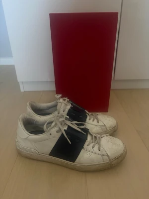 Navy valentinos storlek 45 - Säljer ett par vita sneakers från Valentino med en bred navy rem över snörningen. Skorna har klassisk låg profil, vita skosnören och en chunky sula. Perfekta för dig som gillar stilrena men ändå unika detaljer på dina sneakers.