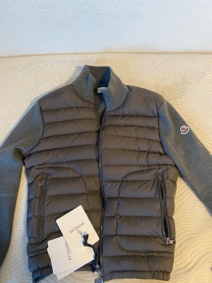 Grå Moncler Cardigan - Snygg grå Cardigan från Moncler med quiltad front och rygg samt stickade ärmar i matchande grått. Västen har två dragkedjeförsedda fickor och hög krage. Moncler-logga på ena ärmen och DIST-certifierad dunfyllning. Perfekt för lager-på-lager och streetstyle.