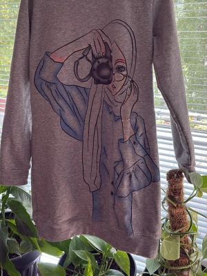 Grå hoodie med konstnärligt tryck - Säljer en grå hoodie med ett stort, konstnärligt tryck av en tjej med kamera på framsidan. Hoodien har långa ärmar och en avslappnad passform. Materialet känns mjukt och bekvämt, perfekt för chill dagar. Ingen synlig logga eller märke.
