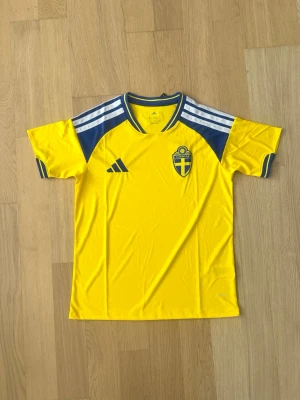 Svensk landslagströja i gult och blått - Gul fotbollströja med blå detaljer och vita ränder på axlarna. Tröjan har Sveriges landslagsemblem på bröstet och är tillverkad i ett lätt, ventilerande material. Perfekt för match eller träning.