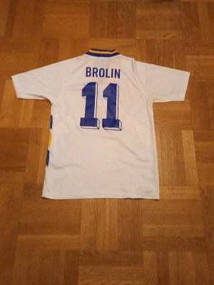 Svensk fotbollströja Brolin #11 Adidas - Retro svensk landslagströja från Adidas med Brolin och nummer 11 på ryggen. Tröjan är vit med blå och gula detaljer, V-ringad krage med blå och gula ränder samt Sveriges landslagsemblem på bröstet. Perfekt för dig som älskar fotboll och klassiska matchtröjor.
