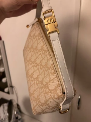 Beige axelremsväska med monogram - Snygg beige handväska med diskret monogrammönster och vita detaljer. Väskan har en justerbar axelrem i vitt läder och guldfärgade metalldetaljer. Perfekt storlek för det viktigaste och stängs med dragkedja upptill.