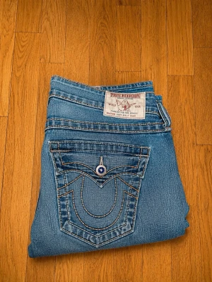 True Religion Joey Bootcut Jeans W27 – Made in USA – Y2K Vintage - Säljer ett par äkta True Religion Joey Bootcut-jeans i storlek W27. Tillverkade i USA med den klassiska Y2K-looken och ikoniska True Religion-detaljer såsom de kraftiga sömmarna och de karakteristiska bakfickorna.  Jeansen är i mycket bra skick och passar perfekt för dig som gillar vintage, 2000-talsmode och bootcut-modellen. Tillverkade i 99 % bomull och 1 % elastan för en bekväm passform.  Mått:  * Midja: 38 cm * Innerbenslängd: 82 cm * Ytterben fram: 100 cm * Ytterben bak: 105 cm