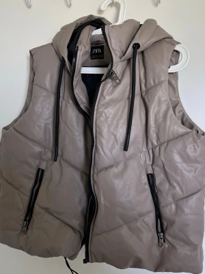 Beige väst med huva från ZARA - Snygg beige dunväst från ZARA med huva och svarta dragskor. Västen har två framfickor med svarta dragkedjor och quiltad design. Perfekt för lager på lager och streetstyle. Materialet är glansigt och ger en modern look.