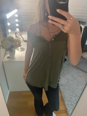Olivgrön ärmlös linnetopp - Snygg olivgrön linnetopp med knappar framtill och avslappnad passform. Toppen är ärmlös och har en lätt v-ringning. Perfekt att matcha med jeans eller kjol för en trendig look.