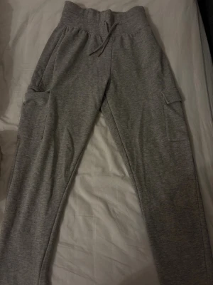 Grå cargo mjukisbyxor med fickor - Säljer ett par grå mjukisbyxor med cargo-inspirerade fickor på sidorna och dragsko i midjan. Byxorna har en avslappnad passform och är gjorda i mjukt sweatshirtmaterial. Perfekta för chill dagar eller när du vill vara bekväm.