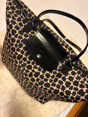 Mönstrad handväska från Longchamp - Snygg handväska från Longchamp med svart, beige och gul grafiskt mönster. Väskan har svarta handtag och lock i glansigt material samt dragkedja. Perfekt för dig som vill ha en unik och rymlig väska med cool design.