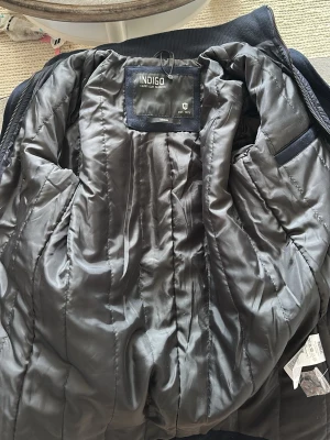 Mörkblå  jacka  från Indigo - Snygg blå jacka från Indigo med quiltad insida och dragkedja. Jackan har en klassisk passform och är fodrad för extra värme. Perfekt för kalla vinterdagar och enkel att matcha med olika outfits.