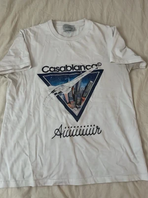 Vit t-shirt från Casablanca - Säljer en vit t-shirt från Casablanca med tryck av ett flygplan och stadsmotiv på bröstet. T-shirten har rund hals och korta ärmar. Trycket har en cool retrovibe och texten 'Aiiiiiiiir' under motivet.