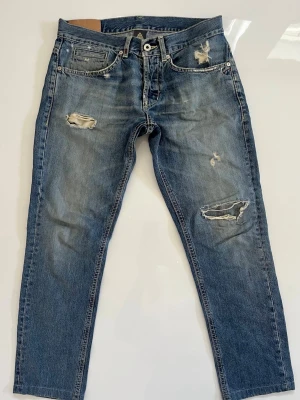 Dondup jeans - Säljer ett par blå jeans med raka ben och flera slitningar på både fram- och baksida. Jeansen har klassisk femficksdesign, knappgylf och är tillverkade i denim. Perfekta för en avslappnad och trendig look.