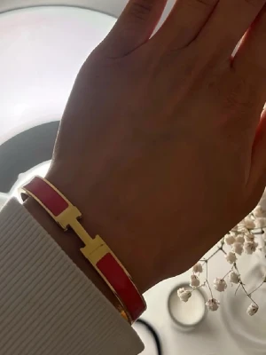 Rött/Orange och guldigt H-armband  - Snyggt armband i röd och guldig metall med en stilren design och ett H-format spänne. Armbandet har en blank finish och känns både trendigt och elegant. Perfekt accessoar för att lyfta din outfit.