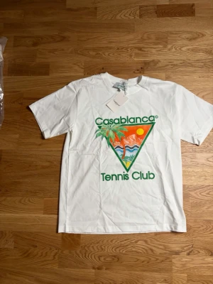 Vit Casablanca Tennis Club t-shirt - Säljer en vit t-shirt från Casablanca med färgglatt tryck på bröstet där det står 'Casablanca Tennis Club' och ett motiv med sol, palmer och berg. T-shirten har rund hals och korta ärmar. Perfekt för dig som gillar sportig och trendig stil.