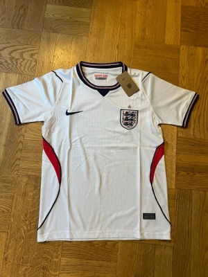 Vit England fotbollströja Nike - Snygg vit England fotbollströja från Nike med broderad landslagssköld och guldfärgad stjärna på bröstet. Tröjan har svarta och röda detaljer längs sidorna och på kragen, samt korta ärmar. Perfekt för match eller träning.