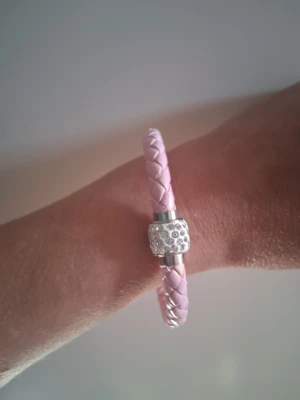 Flätat rosa armband med pärldetalj - Snyggt armband i ljusrosa flätat material med ett silverfärgat lås dekorerat med små glittrande pärlor. Perfekt accessoar för att addera lite extra stil till din outfit. Magnetlås