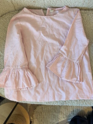 Rosa- och vitrandig blus med volangärm💕 - Söt blus med rosa och vita ränder och volangdetaljer vid ärmsluten. Blusen har rund halsringning och trekvartsärm. Tillverkad i ett lätt och luftigt tyg, perfekt för en fräsch och stilren look.