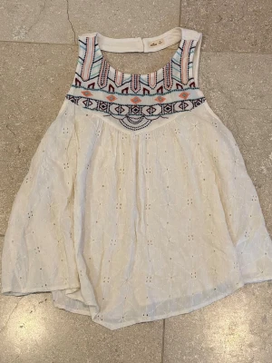 Vit topp med broderat mönster - Luftig vit topp med broderade detaljer och färgglatt etnoinspirerat mönster upptill. Toppen är ärmlös och har en snygg öppen rygg med knappstängning i nacken. Perfekt för varma dagar och ger en bohemisk vibe. Använd flitigt men ser nästan ut som ny:) Broderiet har blivit något slitet genom åren. Säljs i befintligt skick.