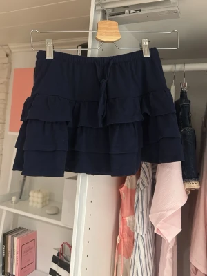 Mörkblå volangkjom från Gina Tricot - Supersöt mörkblå kjol från Gina Tricot med flera lager volanger och snörning i midjan. Perfekta för sommarens alla dagar och ger en lekfull vibe till din outfit. Elastisk midja för extra komfort.