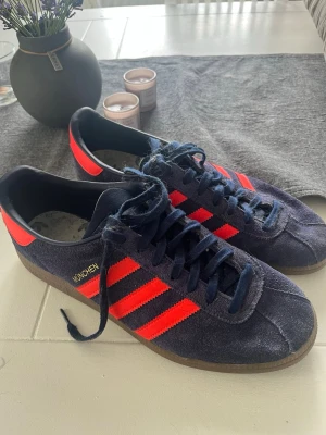 Adidas München blå/röda sneakers - Adidas München sneakers i mörkblå mocka med klassiska röda stripes och detaljer. Låg modell med gummisula och snörning. Snygg retrostil med guldfärgad text på sidan. Perfekta för dig som gillar streetwear och sportig look.