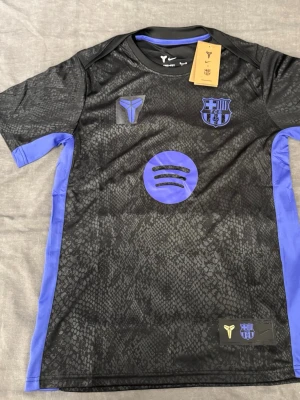 Svart Barcelona x Nike - Säljer en svart Barcelona matchtröja från Nike med lila detaljer och mönstrad yta. Tröjan har FCB-logga, Kobe Bryant-logga och Spotify-logga på bröstet samt lila paneler på sidorna. Tillverkad i Dri-FIT material som andas, perfekt för träning eller gym. Oanvänd helt ny tröja