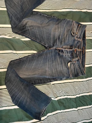Nudie jeans grim tim - Säljer ett par klassiska blå jeans i rak modell med snyggt slitna detaljer och unikt vågmönster på bakfickorna. Jeansen har fem fickor, orangea sömmar och är tillverkade i denim. Perfekta för en avslappnad och trendig look.