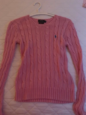 Rosa kabelstickad tröja Polo Ralph Lauren - Säljer en rosa kabelstickad tröja från Polo Ralph Lauren i mjuk pima bomull. Tröjan har rund halsringning, långa ärmar och den klassiska broderade loggan på bröstet. Perfekt för dig som gillar preppy stil och vill ha något bekvämt och snyggt.