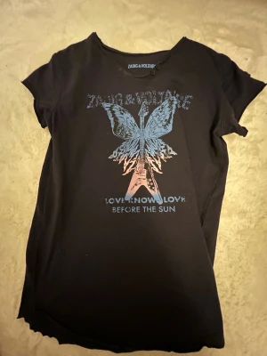 Svart t-shirt med fjärilstryck Zadig & Voltaire - Cool svart t-shirt från Zadig & Voltaire med ett stort tryck av en gitarr med fjärilsvingar i blått och rosa på framsidan. Texten 'LOVE KNOWS LOVE BEFORE THE SUN' under motivet. Klassisk passform och rund hals.