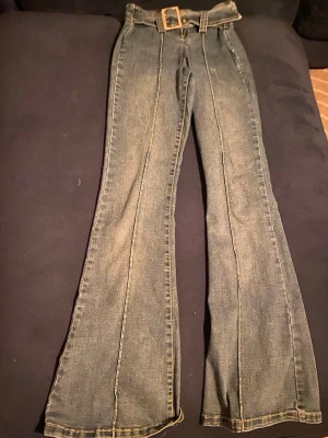 Blå utsvängda jeans med bälte - Säljer ett par blå jeans med mellan midja och utsvängda ben. Jeansen har en bred bältesögla och matchande jeansbälte i midjan. Snygga sömmar framtill ger en retrovibe och de passar perfekt till en trendig vardagslook.