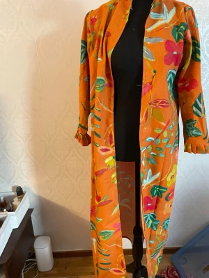 Orange blommig lång kappa - Färgstark lång kappa i orange med stora blommiga mönster i grönt, rosa, rött och blått. Rak passform, öppen framtill utan knäppning och trekvartsärmar. Perfekt statement-plagg för dig som gillar unika och livfulla ytterplagg.