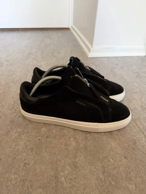 Axel Arigato skor - Snygga svarta sneakers från Axel Arigato med ovandel i mocka och detaljer i läder. Skorna har vit sula och dragkedja framtill istället för klassiska snören. Minimalistisk design med diskret logga på sidan. (Boxen finns ej)