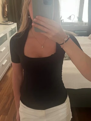 Svart topp med fyrkantig ringning - Snygg svart topp med korta ärmar och en trendig fyrkantig ringning. Modellen är tight och enkel, perfekt att matcha med olika stilar. Materialet ser ut att vara mjukt och stretchigt, vilket ger en bekväm passform.