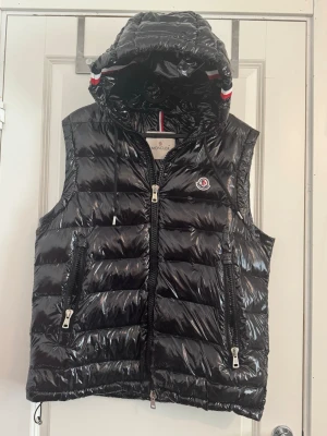 Svart dunväst med huva från Moncler - Snygg svart dunväst från Moncler med glansig finish och avtagbar huva. Västen har två dragkedjefickor fram, Moncler-logga på bröstet och coola detaljer i rött och vitt på huvan. Perfekt för lager-på-lager och streetwear-vibe.