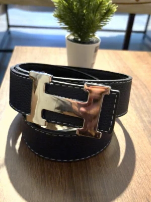 Bälte  - Stilrent svart bälte med ett stort, silverfärgat -format spänne. Bältet har vit kontrastsöm längs kanterna och är tillverkat i skinn. Perfekt accessoar för att lyfta din outfit med en klassisk touch.