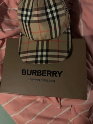 Burberry keps med klassiskt rutmönster - Snygg keps från Burberry i deras ikoniska beigea rutmönster med svarta, vita och röda linjer. Kepsen har böjd skärm och justerbar passform. Perfekt accessoar för att lyfta din outfit med en lyxig touch.