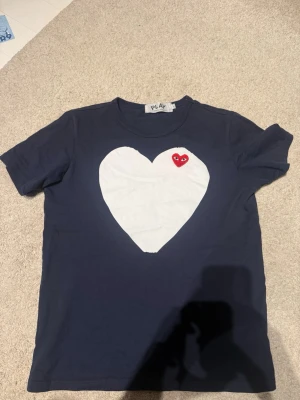Mörkblå t-shirt med hjärta - Säljer en mörkblå t-shirt från come de garçon med ett stort vitt hjärta och ett litet rött hjärta med ögon på bröstet. T-shirten har rund hals och korta ärmar. Märket PLAY syns i nacken. Perfekt för dig som gillar coola prints. Storlek är L men passar bäst på M