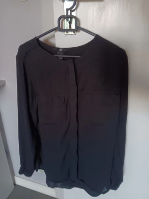 Svart blus med fickor från H&M - Stilren svart blus från H&M med rund halsringning och två bröstfickor. Blusen har långa ärmar och är tillverkad i ett tunt, lätt material som ger ett snyggt fall. Perfekt att matcha med jeans eller kjol för en clean look.  Blusen är använd en gång . 