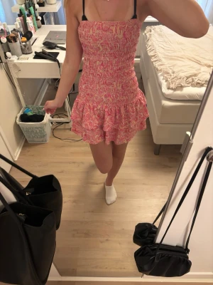 Rosa mönstrad kort klänning - Supersöt kort klänning i rosa med volangkant och smockad överdel. Klänningen har ett blommigt mönster i rosa, vitt och gult och är axelbandslös. Perfekt för varma dagar och ger en somrig vibe.