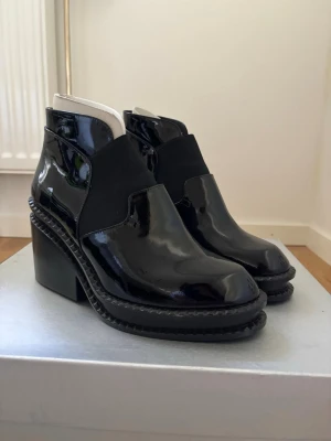 Robert Clergerie ankle boots - Snygga svarta stövletter i glansigt lackmaterial med bred klack och markerad sula. Skorna har en stilren design med elastiska partier på sidorna för enkel påtagning. Perfekta för att lyfta din outfit med en edgy touch.