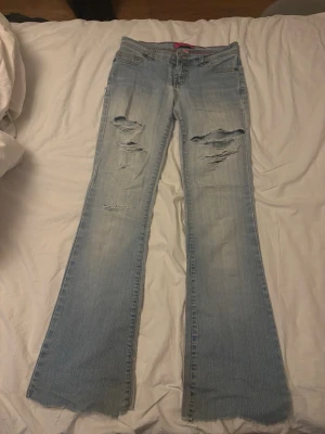 Ljusblå slitna bootcut jeans - Säljer ett par ljusblå bootcut jeans med slitningar och hål på både fram- och baksida. Jeansen har råa, fransiga benslut och klassisk femficksmodell. Perfekta för en avslappnad och trendig look. Jensen har ett hål vid rumpan på baksidan.