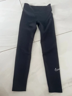 Svarta Nike träningstights - Svarta träningstights från Nike med vit swoosh-logga på ena benet. Tightsen har hög midja och är tillverkade i ett stretchigt material som sitter tajt mot kroppen. Perfekta för gym, löpning eller annan sport.