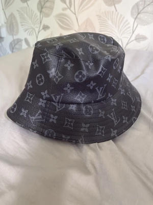 Svart bucket hat med monogram - Snygg svart bucket hat i läderimitation med grått monogrammönster över hela hatten. Klassisk form med mjuk brätte och fodrad insida. Perfekt accessoar för att lyfta din streetwear-look.