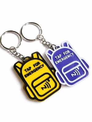 NFC-tagg till barn - Två nyckelringar i form av ryggsäckar, en gul med svart text och en lila med vit text. Båda har texten 'Tap for Emergency' och en symbol för kontaktlös. Tillverkade i plast med metallkedja och ring. Med denna nyckelring kan någon bara hålla sin telefon mot nyckelringen och då ringer den automatiskt upp ett valfritt nummer. Perfekt som en extra trygghet för barn eller äldre. 