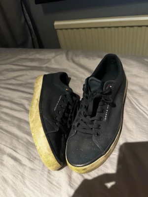 Svarta Tommy Hilfiger canvas sneakers - Snygga svarta sneakers från Tommy Hilfiger med klassisk låg profil och vita sulor. Skorna har textilöverdel, svarta skosnören och diskret logga på sidan. Perfekta för en clean och stilren look.   Lite gröna men bara efter jag klippt gräs i dom tvättar bort de innan köp såklart dom är i väldigt väldigt bra skick efter man tvättat dom nästan nya 